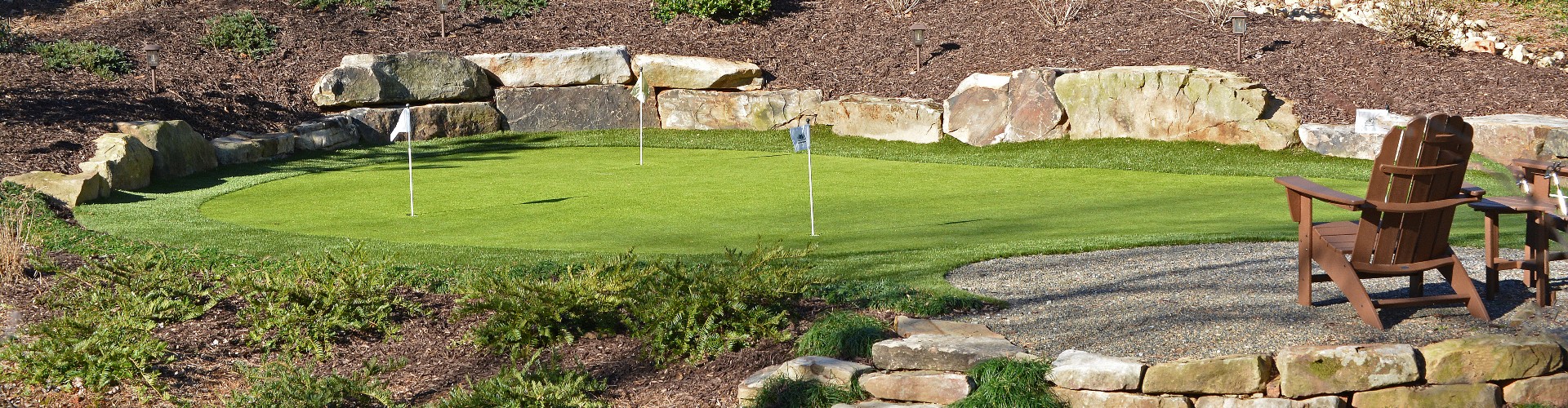Southwest Greens backyard putting green with two flag holes, natural boulder landscaping, and Adirondack seating area.